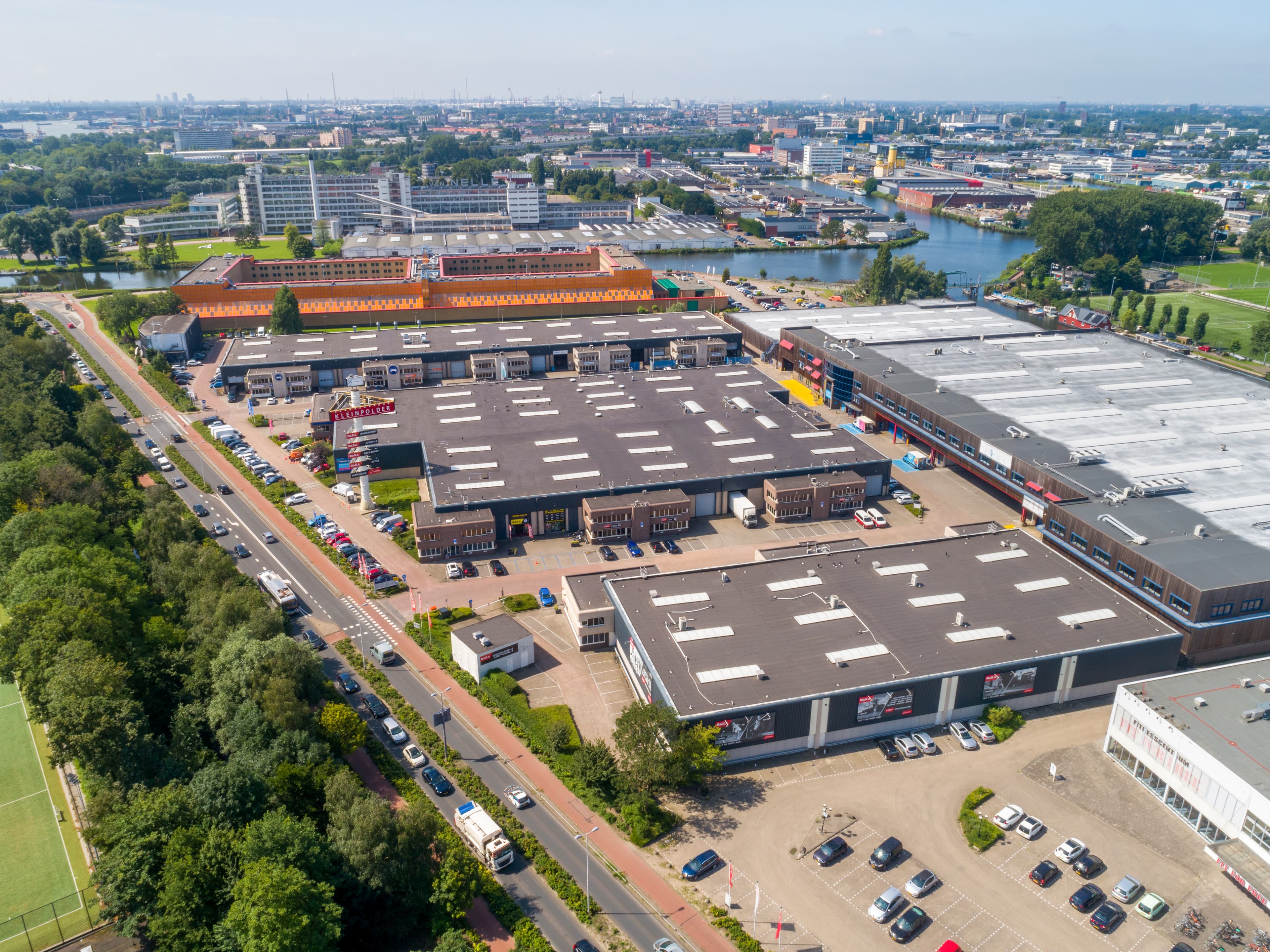 Aerial view of an industrial estate featuring multiple large warehouses, car parks, surrounding roads, and areas of greenery under a clear sky.