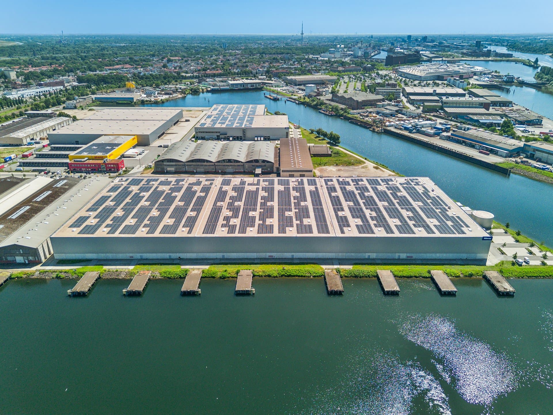 Aerial view of large industrial warehouses with solar panels on the roofs, located next to a canal in an urban area.