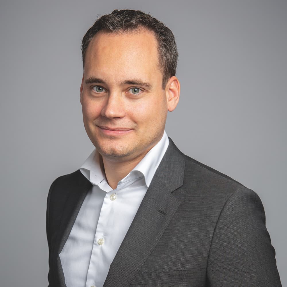 A person in a dark suit and white shirt stands in front of a plain grey background, looking at the camera with a slight smile.