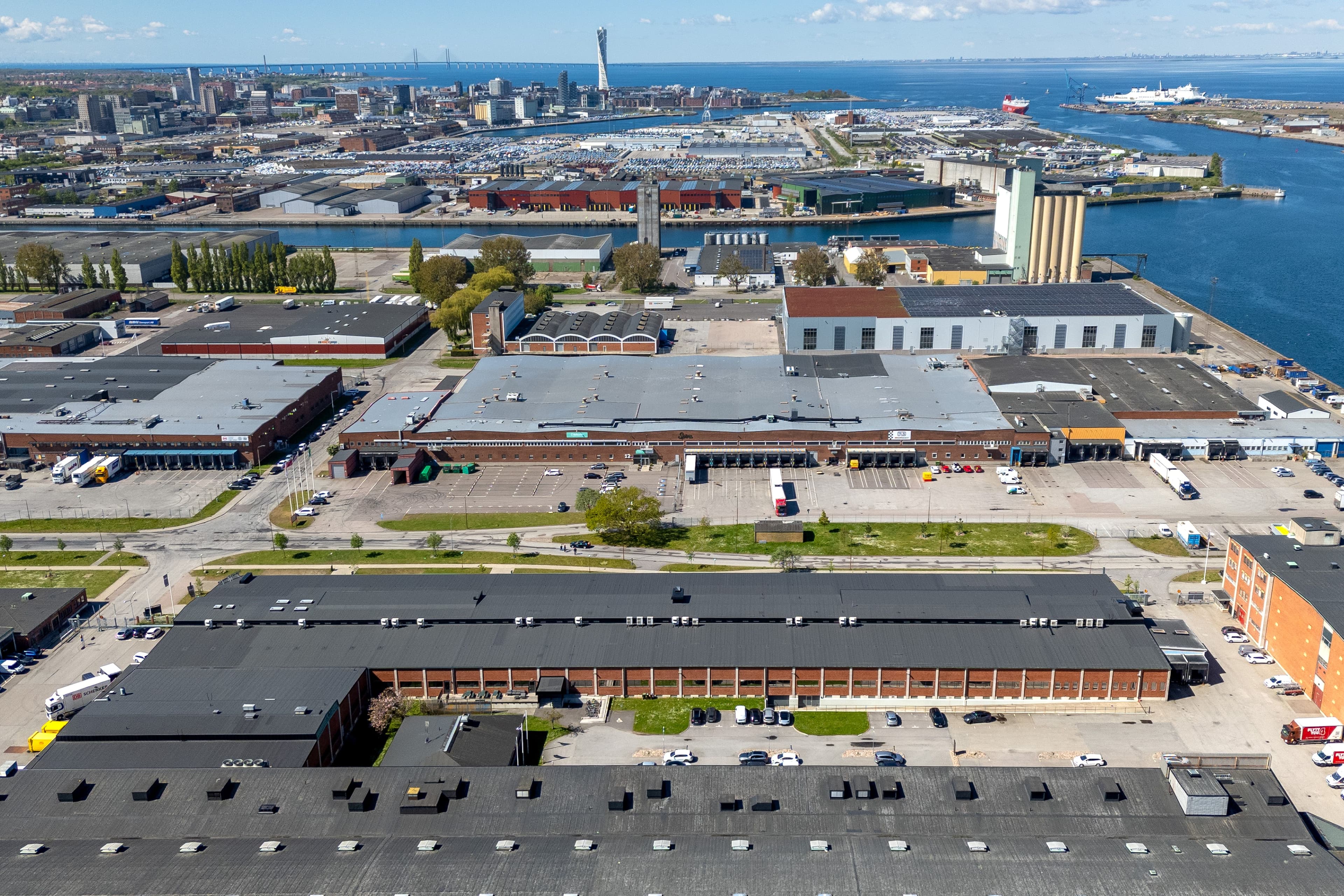 Aerial view of an industrial area near a harbour, featuring warehouses, parked vehicles, water, and a city skyline in the background under a partly cloudy sky.