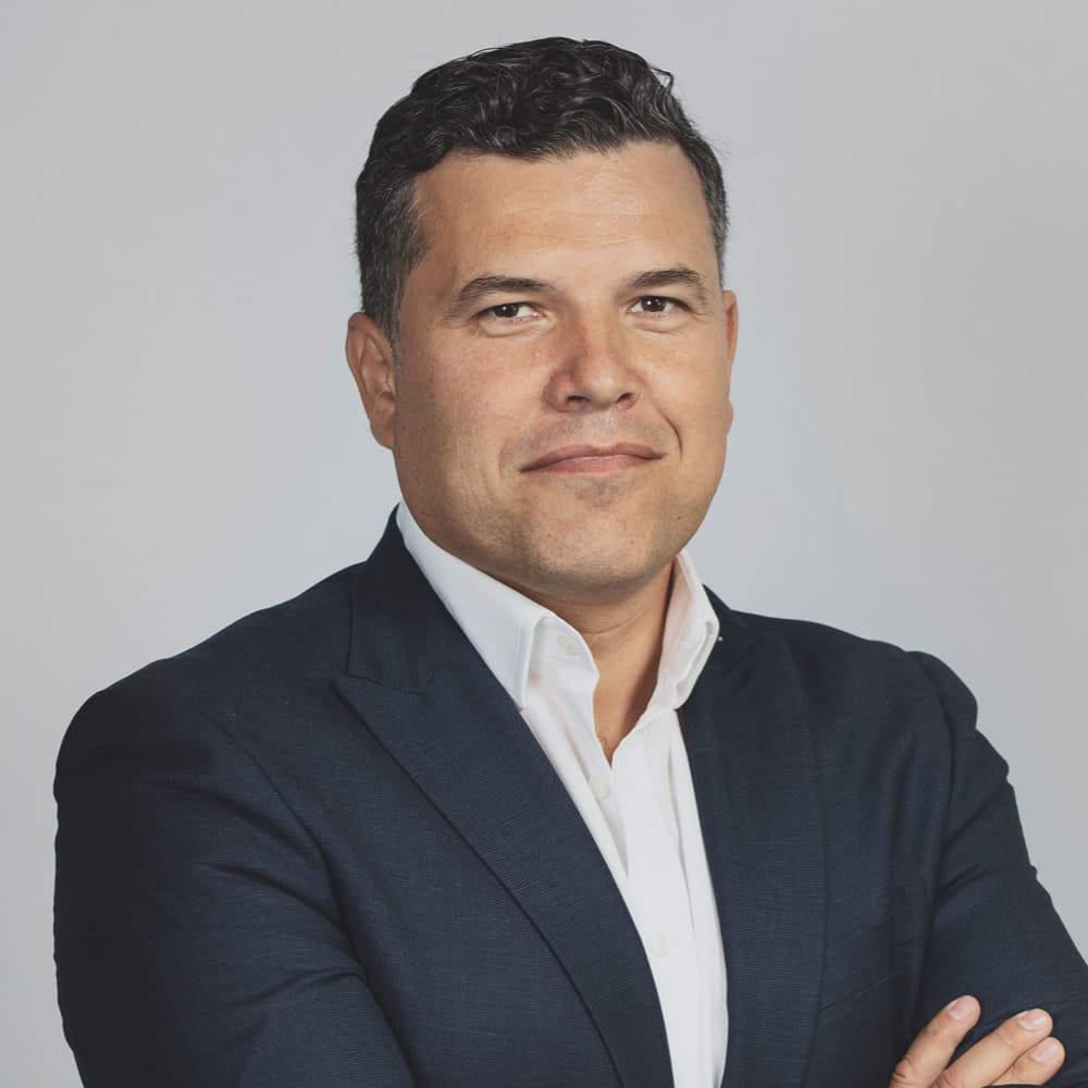 Person in a navy suit with a white shirt standing against a plain light background, arms folded and facing the camera with a neutral expression.