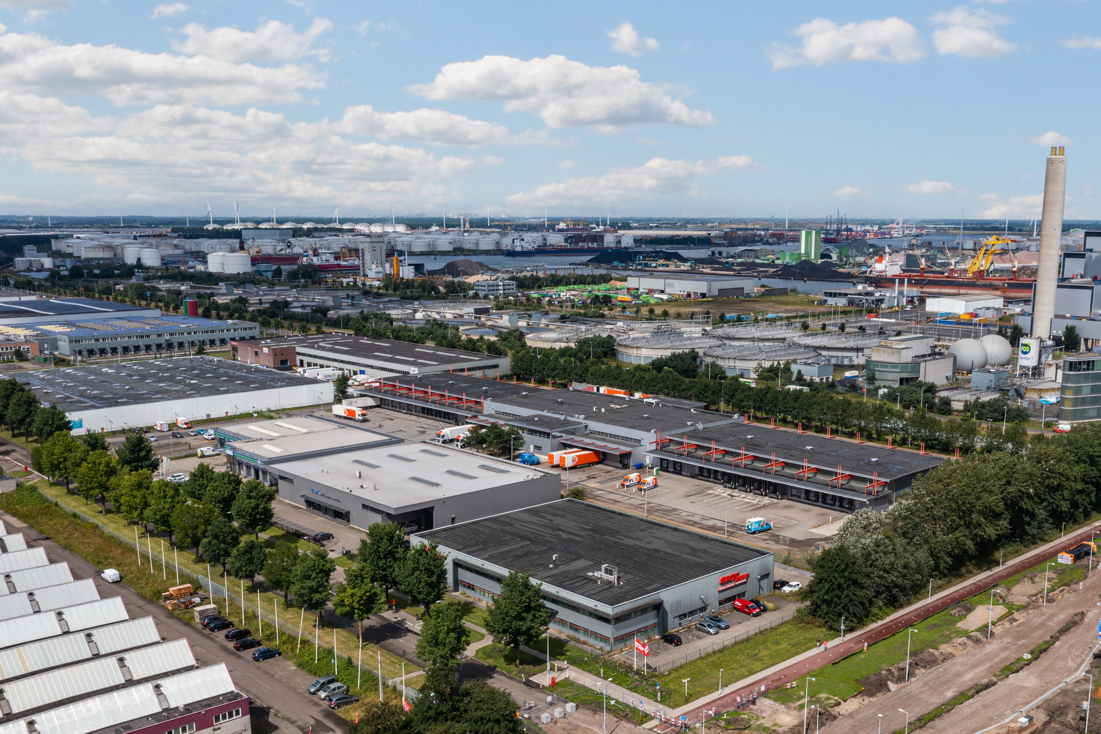 Aerial view of an industrial area featuring warehouses, factories, car parks, and green spaces under a partly cloudy sky.
