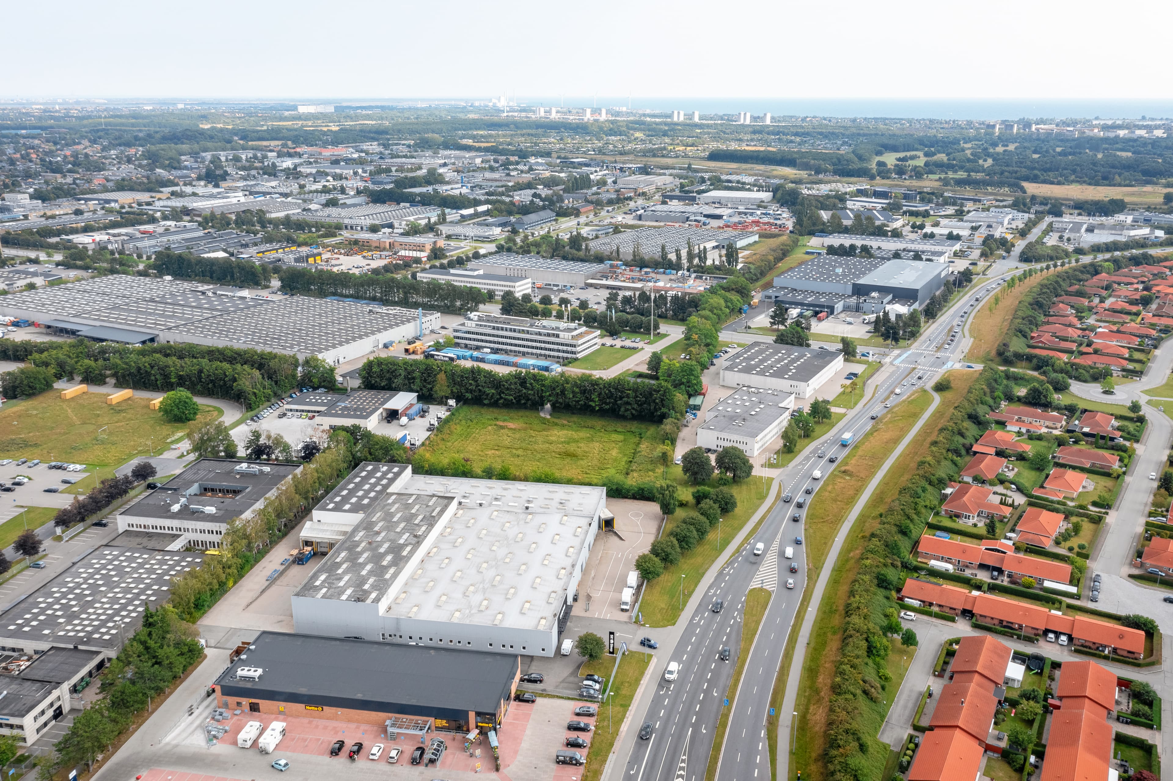 Aerial view of an industrial area with warehouses, roads, and nearby residential houses, featuring traffic and greenery throughout the landscape.