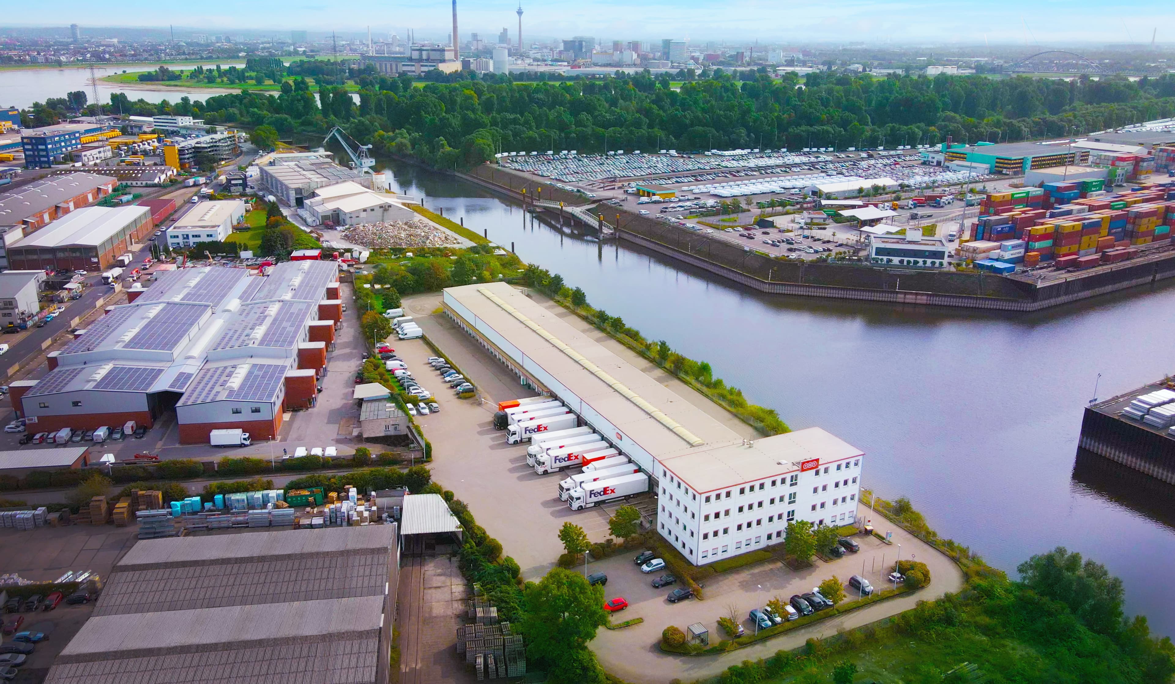Aerial view of an industrial area with warehouses, parked lorries, shipping containers, a river, and a city skyline in the background beneath a cloudy sky.