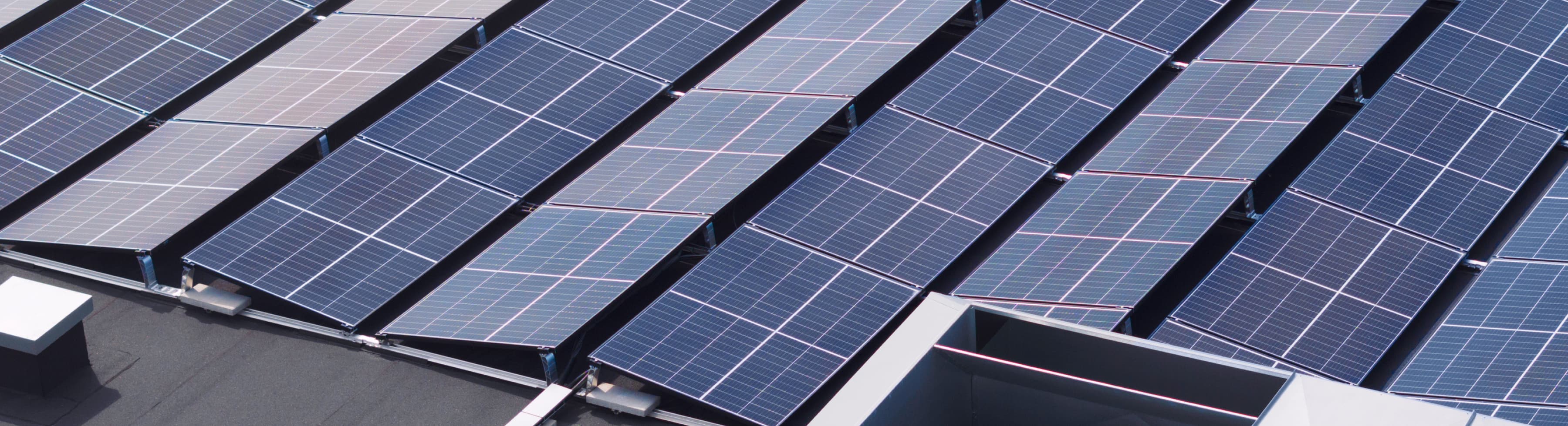 Rows of solar panels installed on a rooftop are shown in sunlight, viewed from above.