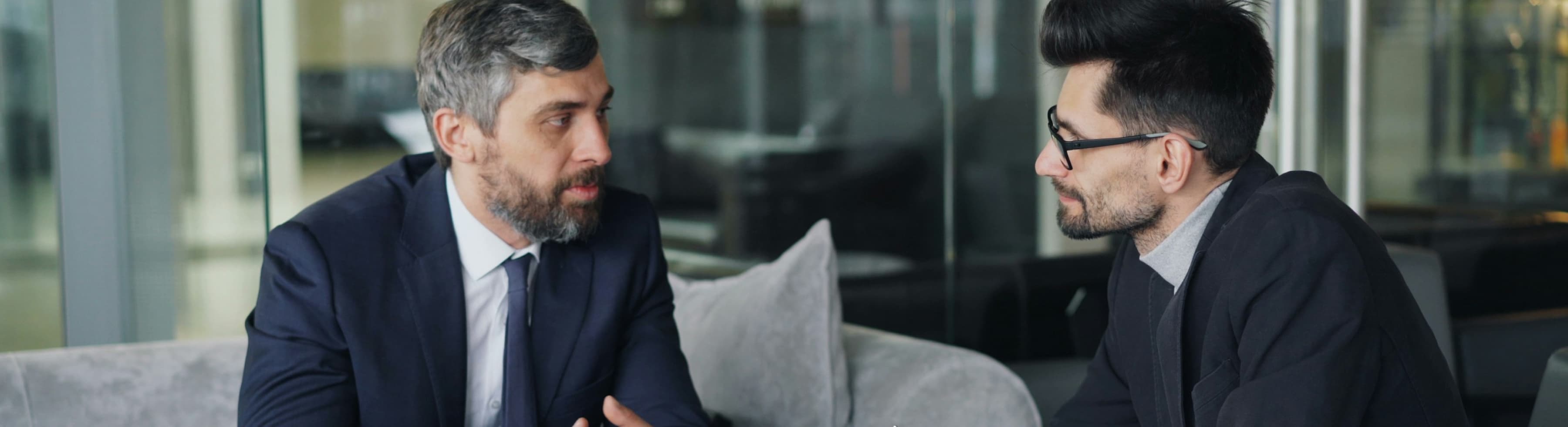 People in business attire are sitting on a sofa in an office, engaged in a serious conversation.
