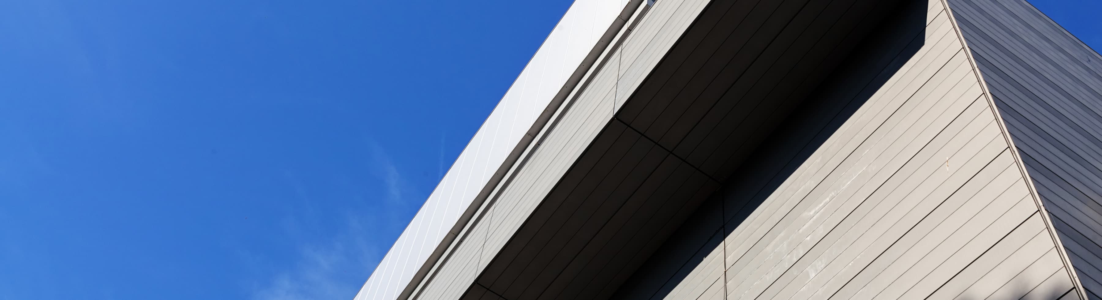 A close-up view of a modern building’s corner features beige panels and a sharp geometric overhang against a clear blue sky.