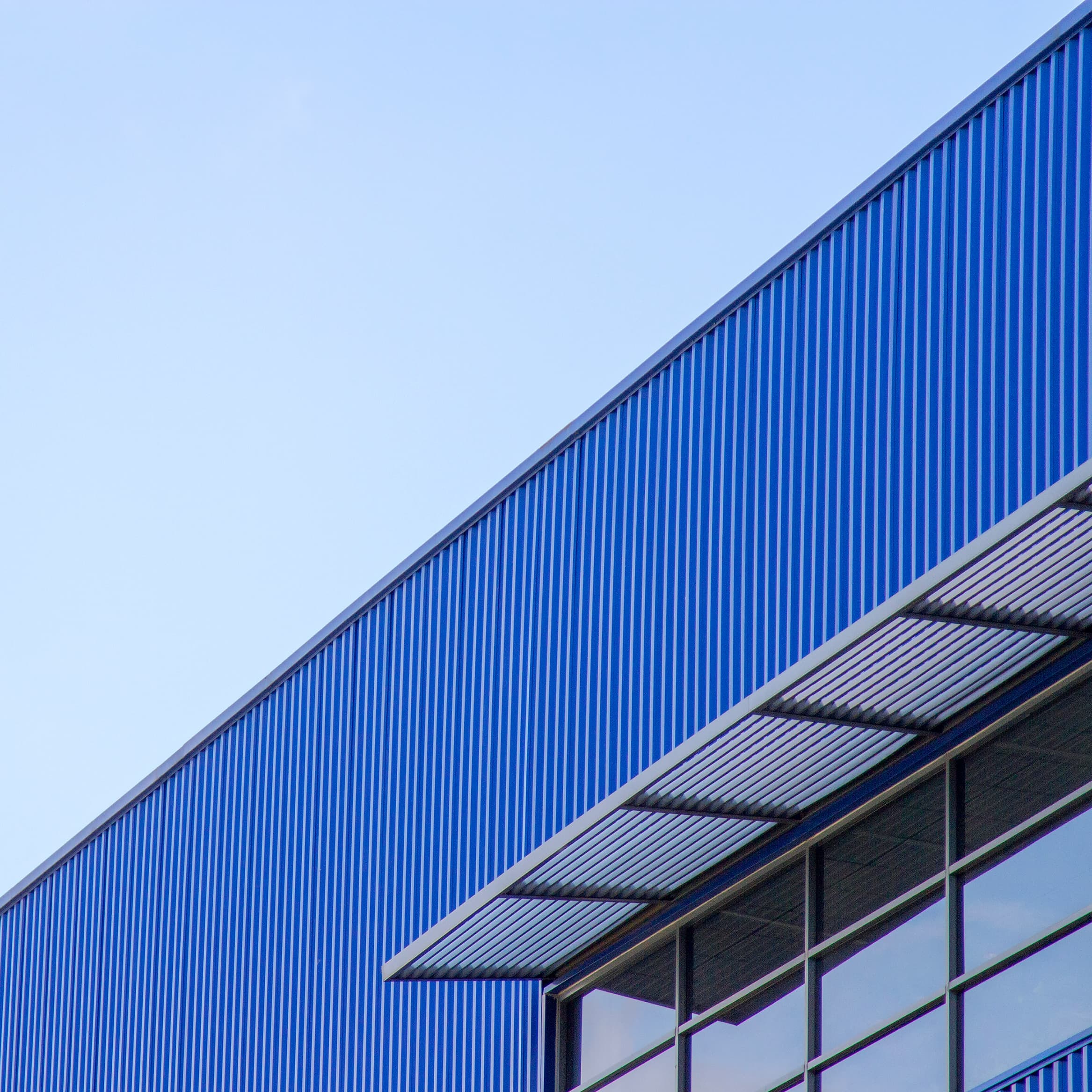 Exterior view of a modern industrial building with blue vertical metal cladding and large windows, under a clear sky.